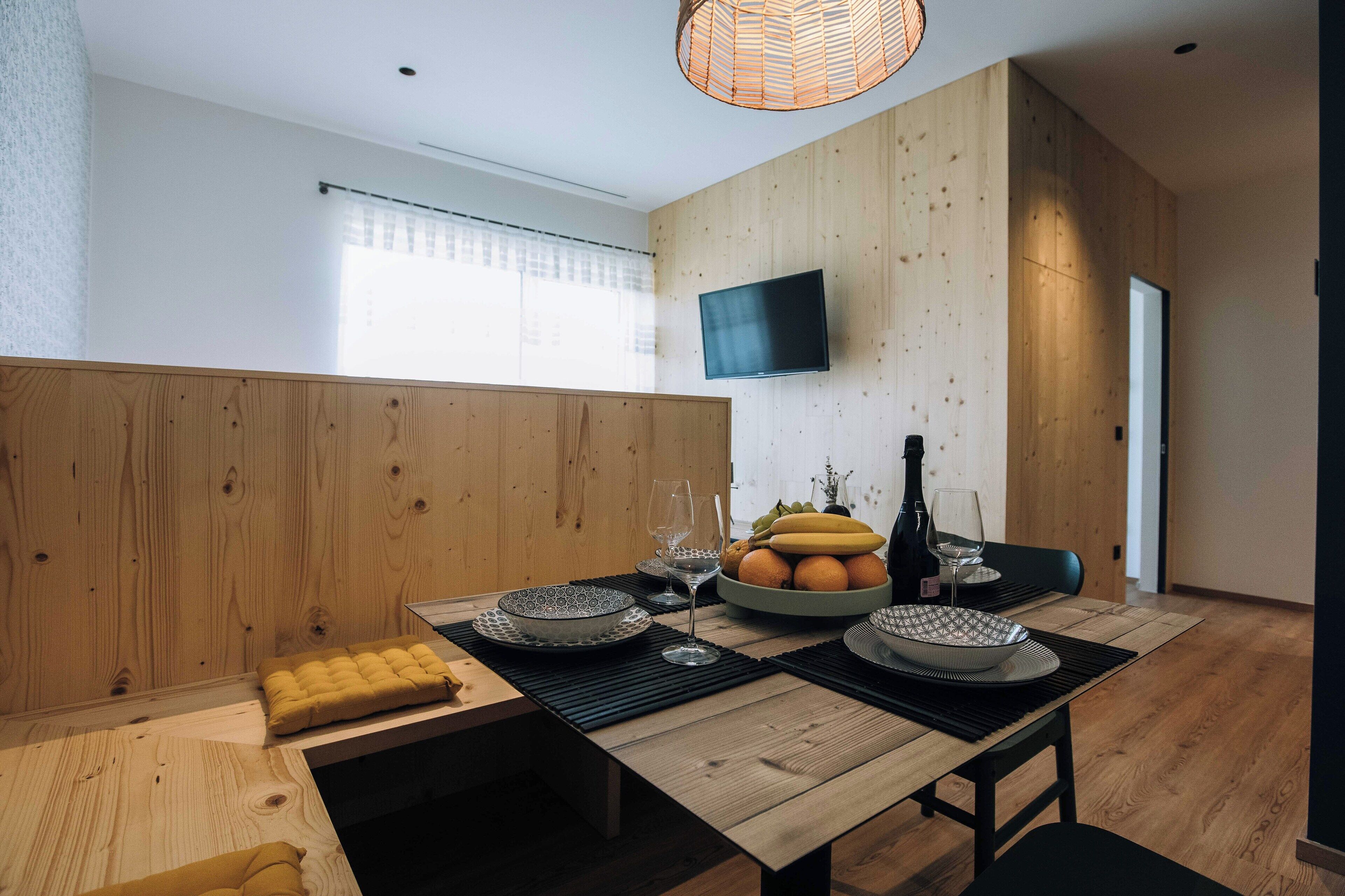 A modern dining area with a wooden table set for two, a television mounted on the wall, and a window with blinds.