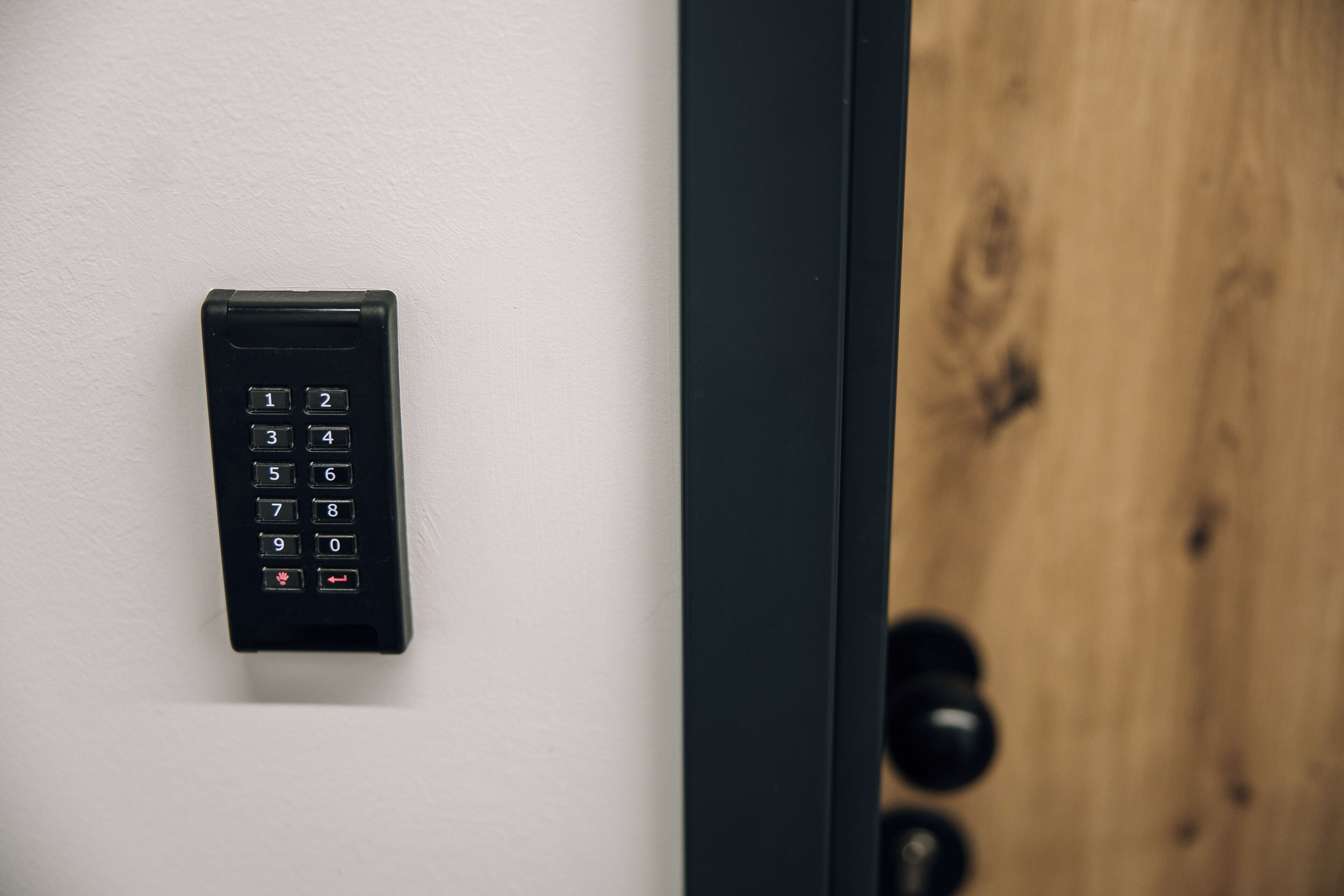A keypad lock on a white wall, next to a wooden door with a black handle.