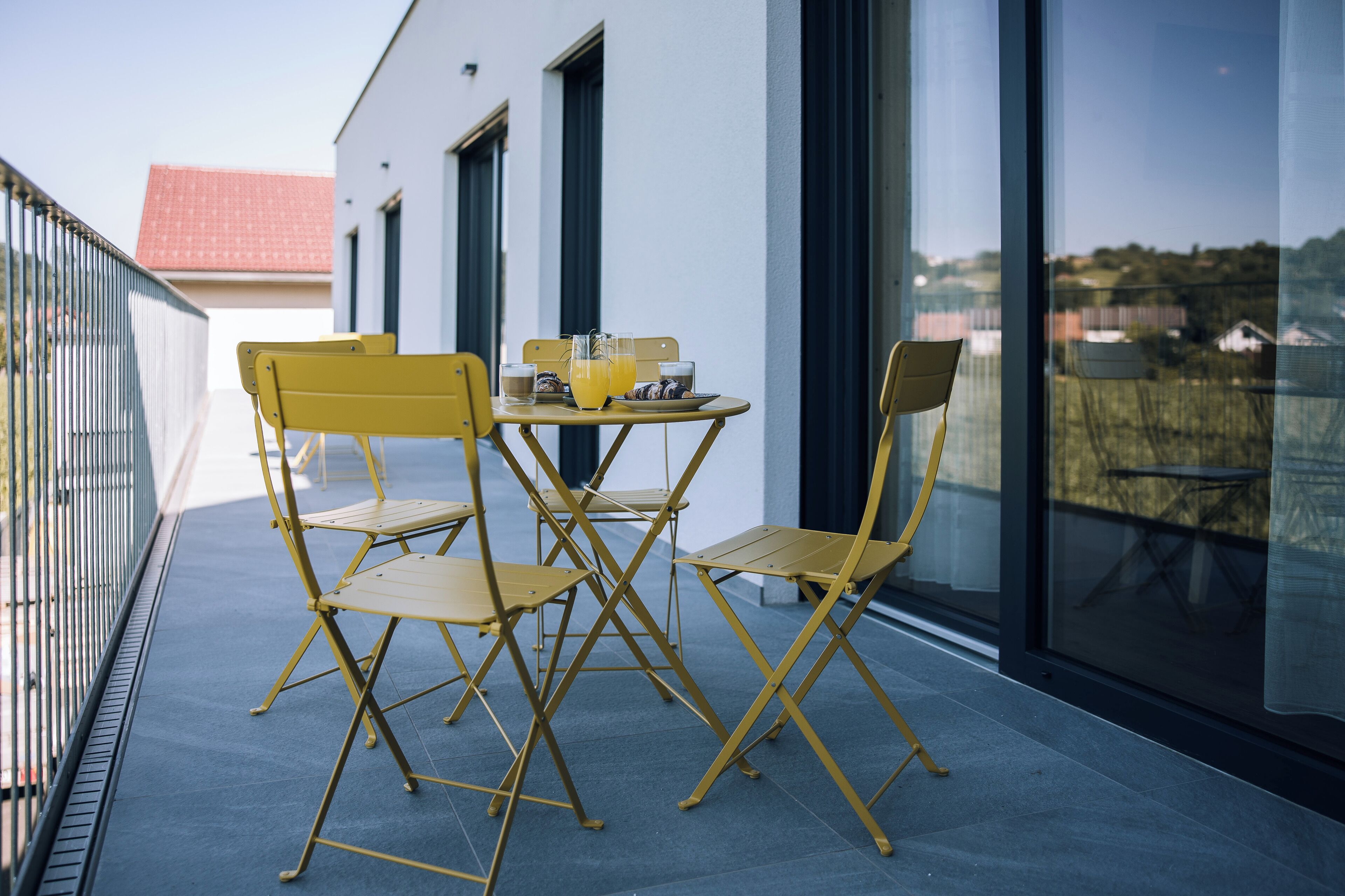 A balcony with yellow chairs and a table, a glass door, and a view of a building with a red roof.
