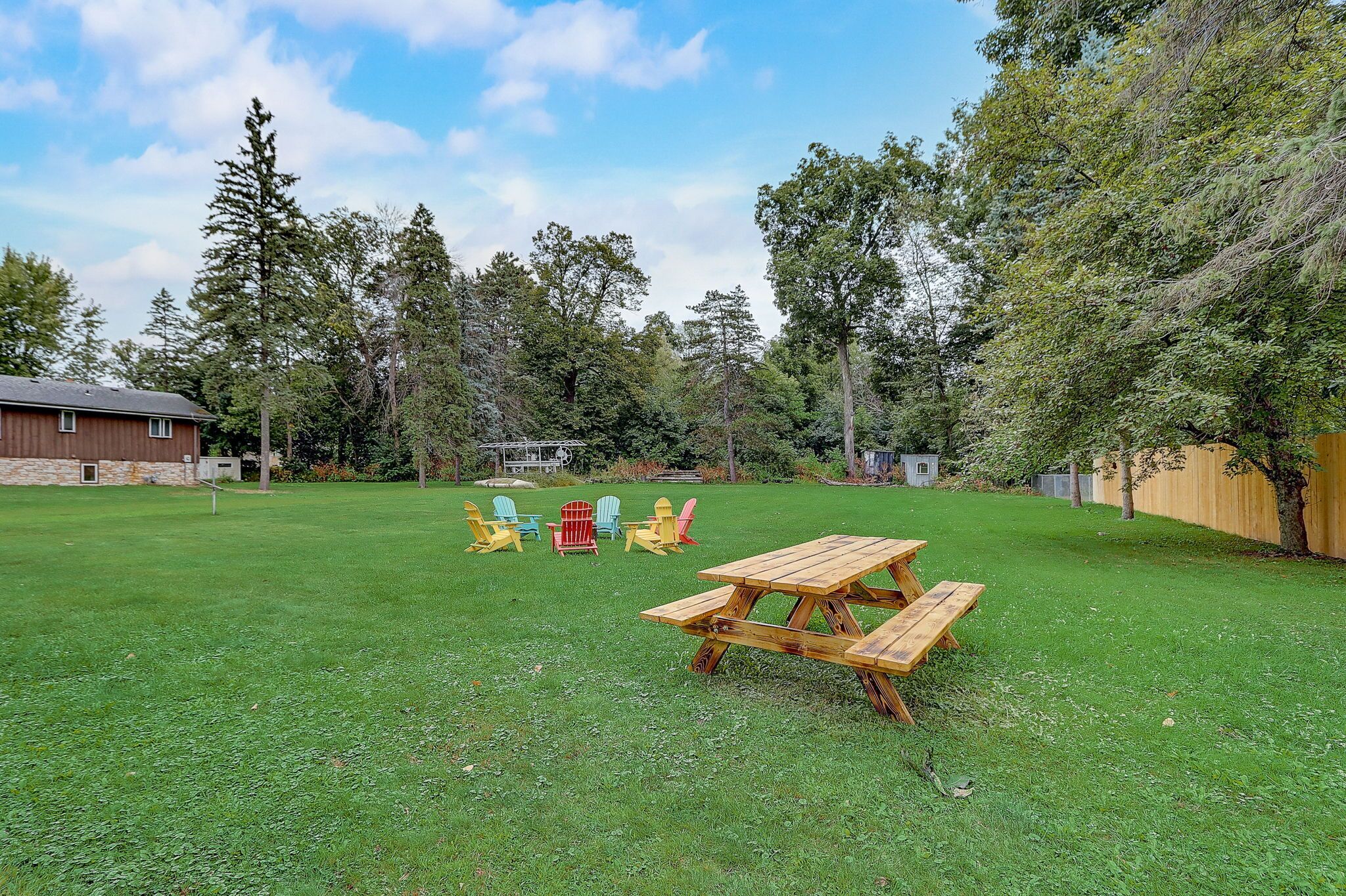 Un patio trasero con una mesa de picnic de madera y sillas Adirondack de colores.
