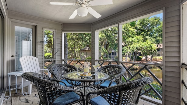 A covered patio with wicker chairs and a table, overlooking a body of water and trees.