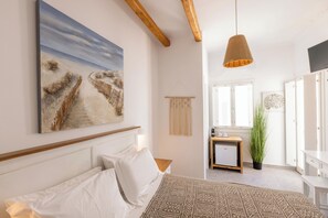A bedroom with a bed, a painting, a hanging light fixture, and a view of a hallway. - Parosquare Central Suites (Paros)