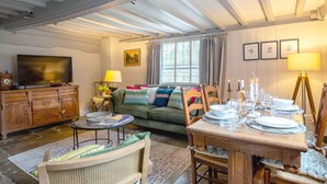 A cozy living room with a television, a sofa, a dining table set for four, and a window with curtains. - Locke's Cottage - sleeps 2 guests  in 1 bedroom (Stratford-upon-Avon)