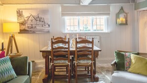 A dining area with a wooden table set for four, flanked by two chairs with cushioned seats. A window with white curtains allows natural light to enter, and a painting of a house hangs on the wall. - Locke's Cottage - sleeps 2 guests  in 1 bedroom (Stratford-upon-Avon)