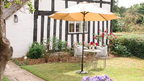 A garden with a table and chairs under a yellow umbrella, surrounded by blooming flowers and a white building with black trim. - Locke's Cottage - sleeps 2 guests  in 1 bedroom (Stratford-upon-Avon)