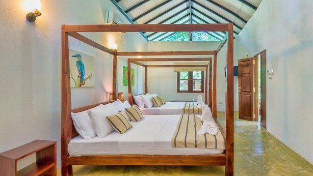 A large wooden four-poster bed with multiple pillows and a blanket, a wooden nightstand, and a painting of a bird on the wall. - Thuduwa Eco Resort  (Rankethgama)