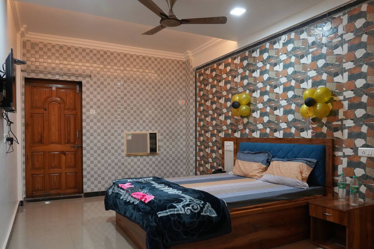 A bedroom with a checkered wall, a wooden door, a ceiling fan, a bed with a blue headboard, and decorative balloons.