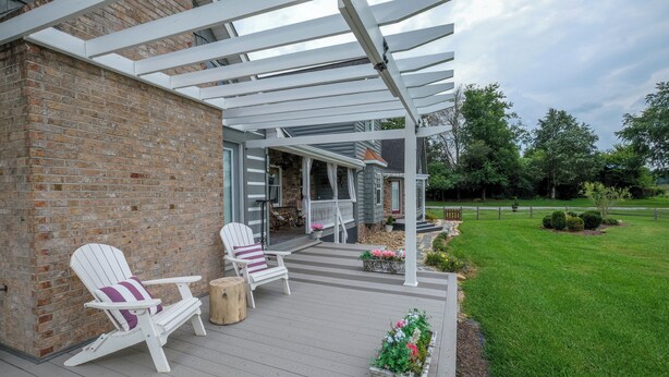 Een overdekt terras met twee Adirondack-stoelen, een houten krukje en een bloemenplanter.