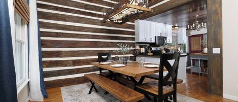 A dining area with a wooden table and benches, a hanging light fixture, and a kitchen in the background.