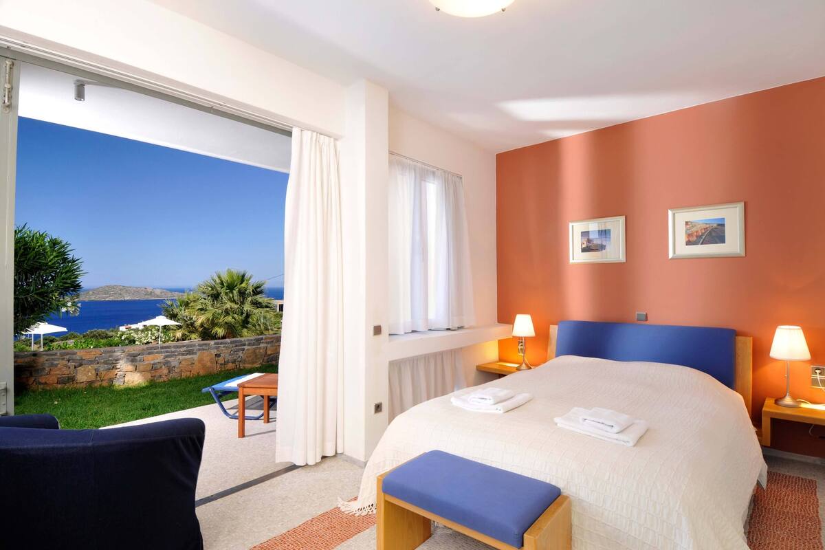 Une chambre d’hôtel avec un lit, deux tables de chevet avec des lampes, une fenêtre avec des rideaux et une vue sur la mer.