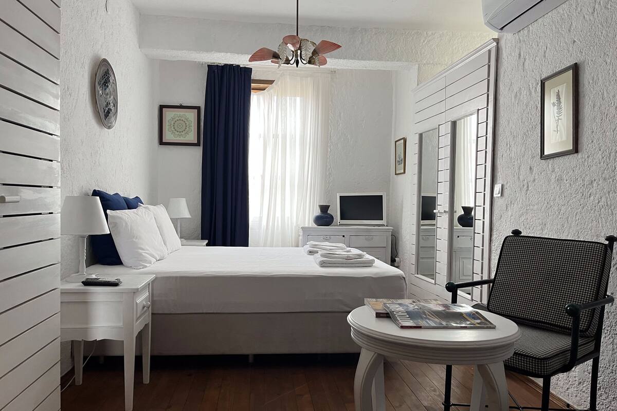 Ein Hotelzimmer mit Bett, Nachttischen, einem Sessel, einem kleinen Tisch, einem Fernseher und einem Fenster mit Vorhängen.