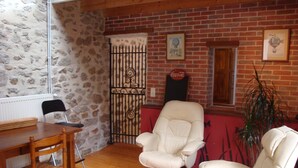 A room with a stone wall, a brick fireplace, a white leather armchair, a wooden dining table with chairs, and a potted plant.