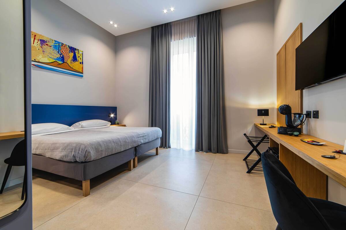 A modern hotel room with a bed, desk, chair, and a large window with curtains.