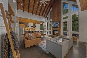 A spacious living area with a fireplace, a kitchen in the background, and large windows.