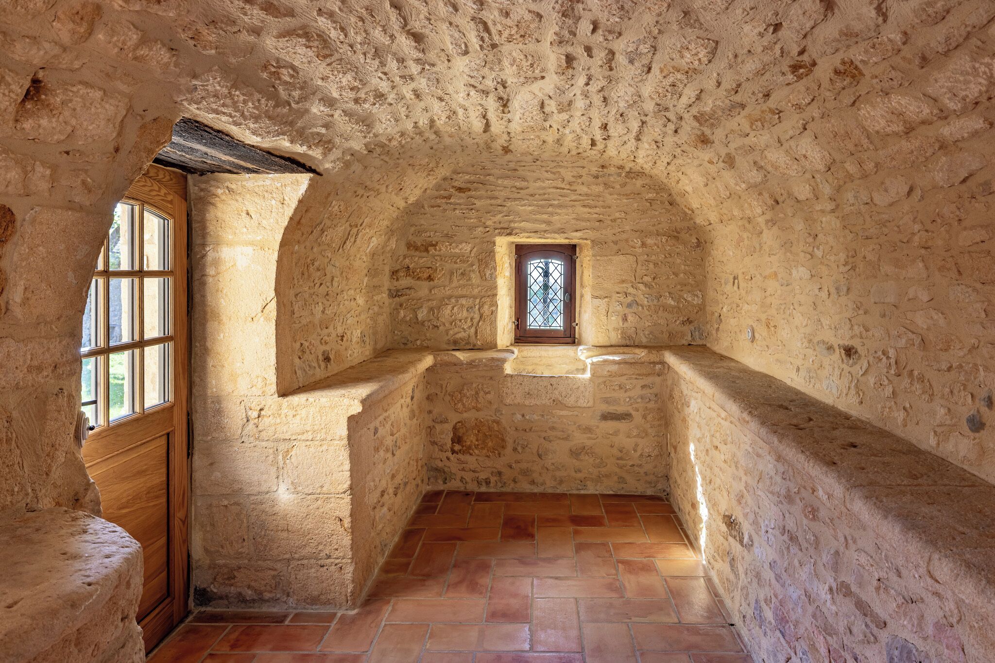 A stone-walled room with a wooden door, a small window, and a tiled floor.