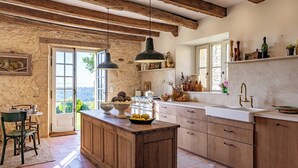 A rustic kitchen with a wooden island, stone walls, and a large window. - Promo Dernière Minute Magnifique (Beynac-et-Cazenac)