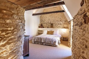 Une chambre avec des murs en pierre, un plafond en bois, un lit recouvert d’une courtepointe beige et une table de chevet avec une lampe.