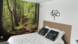 Un dormitorio con un mural grande de un bosque, una cama con cubrecama de estampado geométrico, dos almohadas estampadas, un cabecero de madera y una mesita de noche con una lámpara.