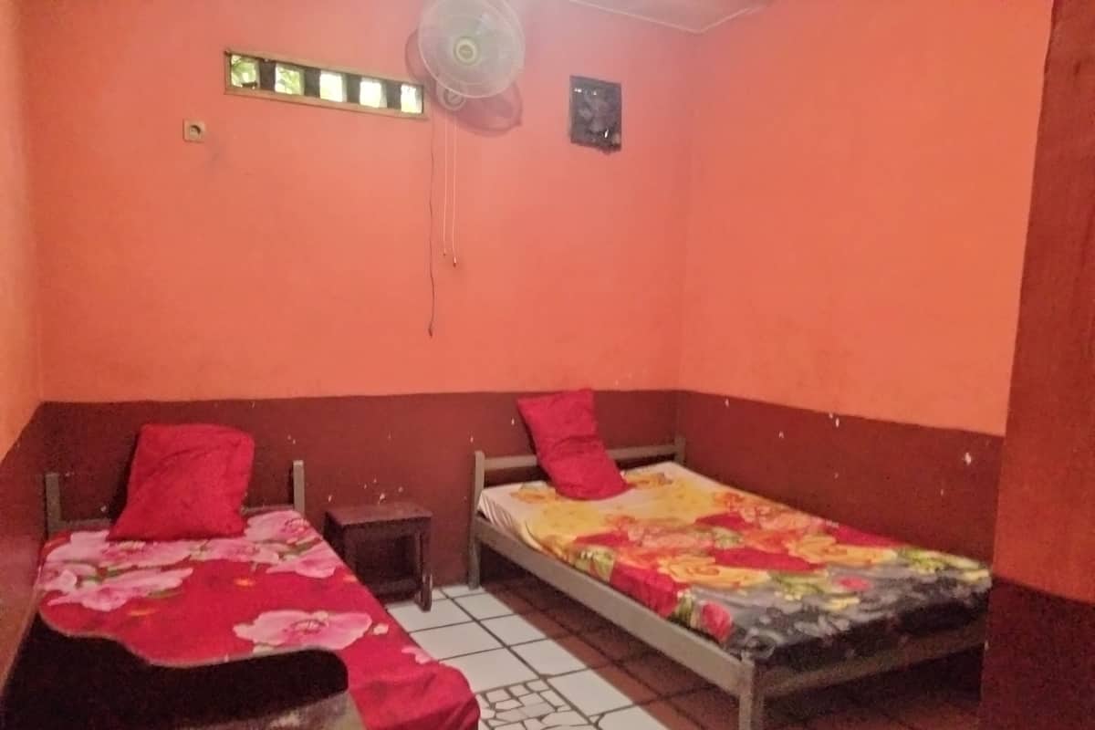 A room with two beds, each with a red pillow and a floral bedspread. There is a small wooden nightstand between the beds, and a ceiling fan above.