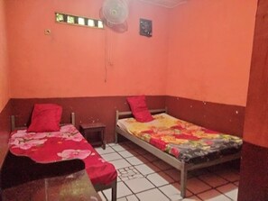 A room with two beds, each with a red pillow and a floral bedspread. There is a small wooden nightstand between the beds, and a ceiling fan above.