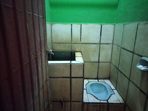 A small, tiled bathroom with a squat toilet, a faucet, and a shelf.