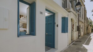 A narrow street with a white building featuring a blue door and shutters, a visible staircase, and a number '17' sign.