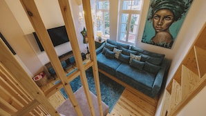 A cozy living room with a blue sofa, wooden beams, a painting, and a view through a window.
