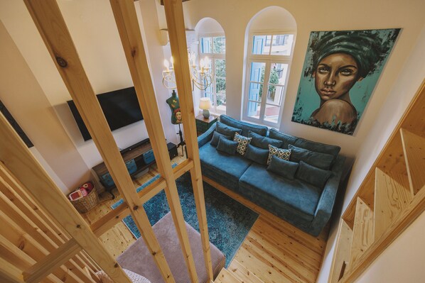 A cozy living room with a blue sofa, wooden beams, a painting, and a view through a window.