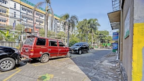 Een straat met geparkeerde voertuigen, waaronder een rode bestelwagen en een zwarte auto. Op de achtergrond staan gebouwen, waarvan er één een bord heeft met de tekst 'POLITEKNIK PELASA'.