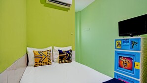Een bed met witte beddengoed en twee kussens met pijlpuntmotieven, een televisie bevestigd boven een blauwe kast met lades, en een airconditioningunit aan de muur.