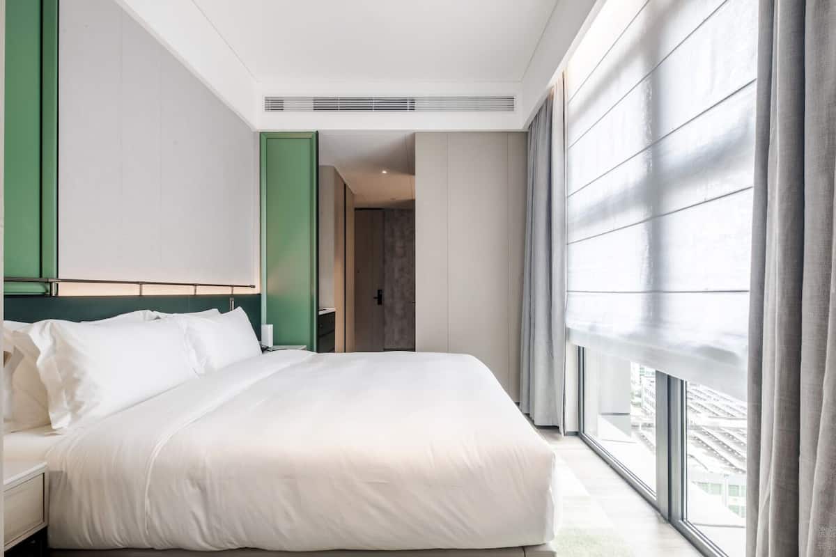 A modern hotel room with a large bed, a view of the city through floor-to-ceiling windows, and a green accent wall.