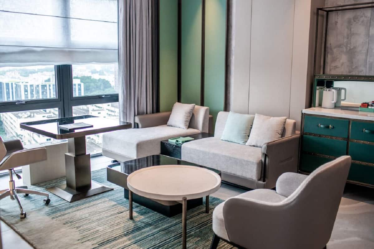 A modern hotel room with a sofa set, a round coffee table, a desk with a chair, and a view of the city through large windows.