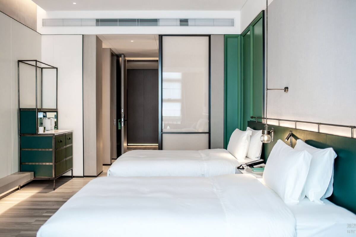 A modern hotel room with two beds, a green accent wall, a large mirror, and a desk with a lamp.