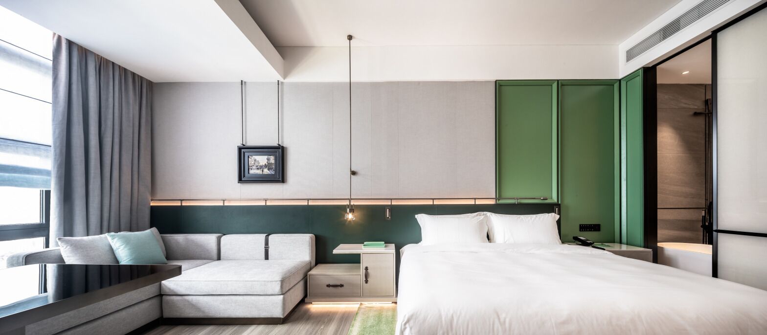 A modern hotel room with a bed, a sofa, a nightstand, and a desk.