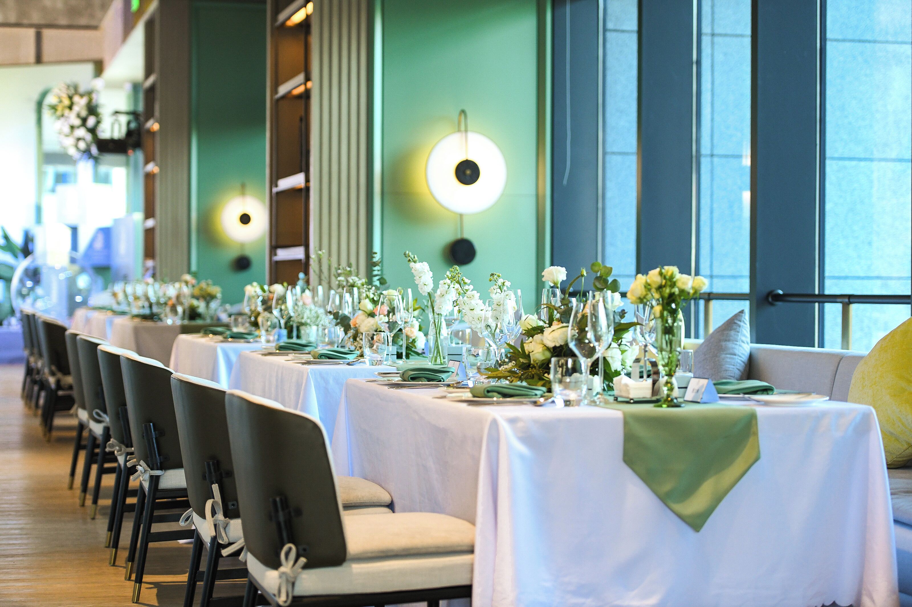 A long dining table with chairs, set with white tablecloths and floral centerpieces.