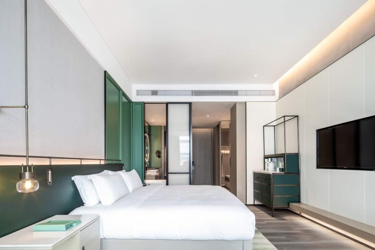 A modern hotel room with a large bed, a bedside table, a TV mounted on the wall, and a walk-in closet.