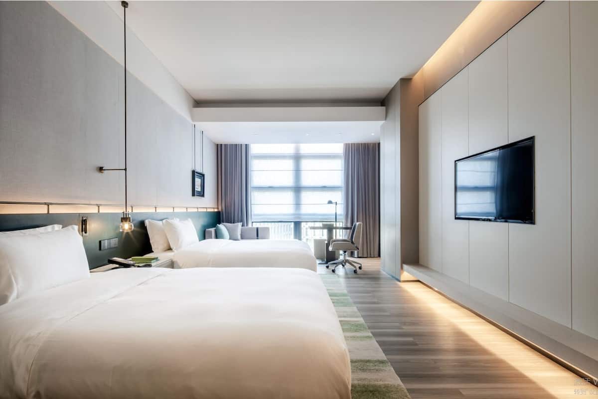 A modern hotel room with two large beds, a flat-screen TV mounted on the wall, and a view of the city through large windows.