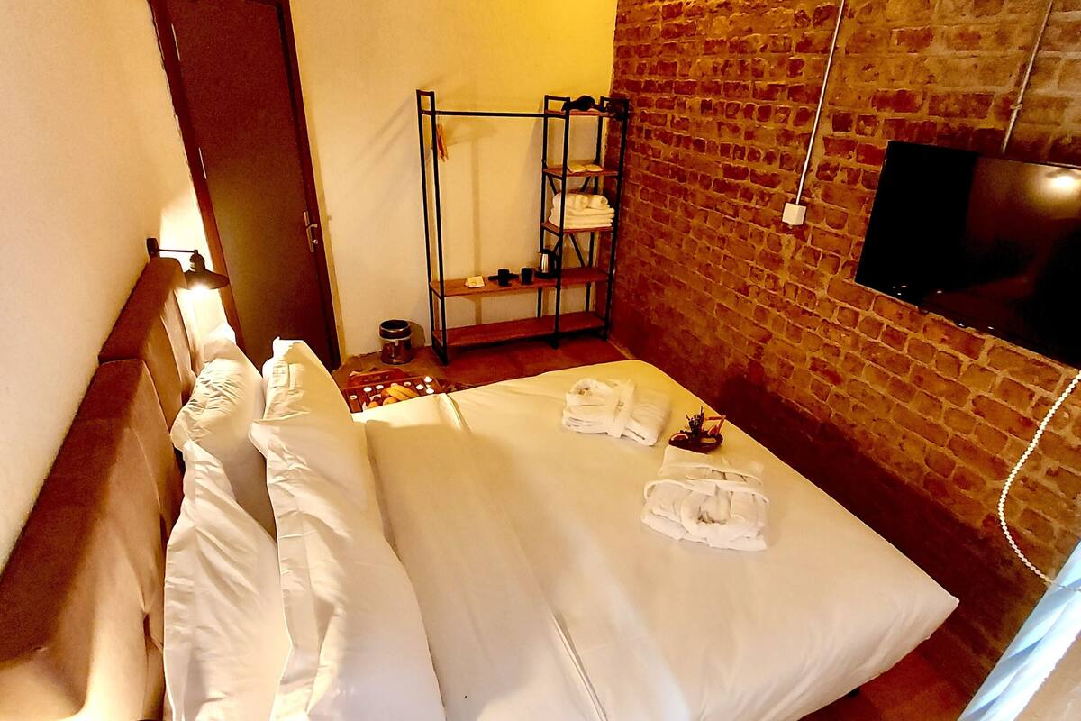 A hotel room with a brick wall, a bed with white linens, a shelf with towels, a TV mounted on the wall, and a door.