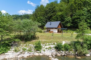 A cabin with a sloped roof, surrounded by a natural landscape with a stream and dense trees.