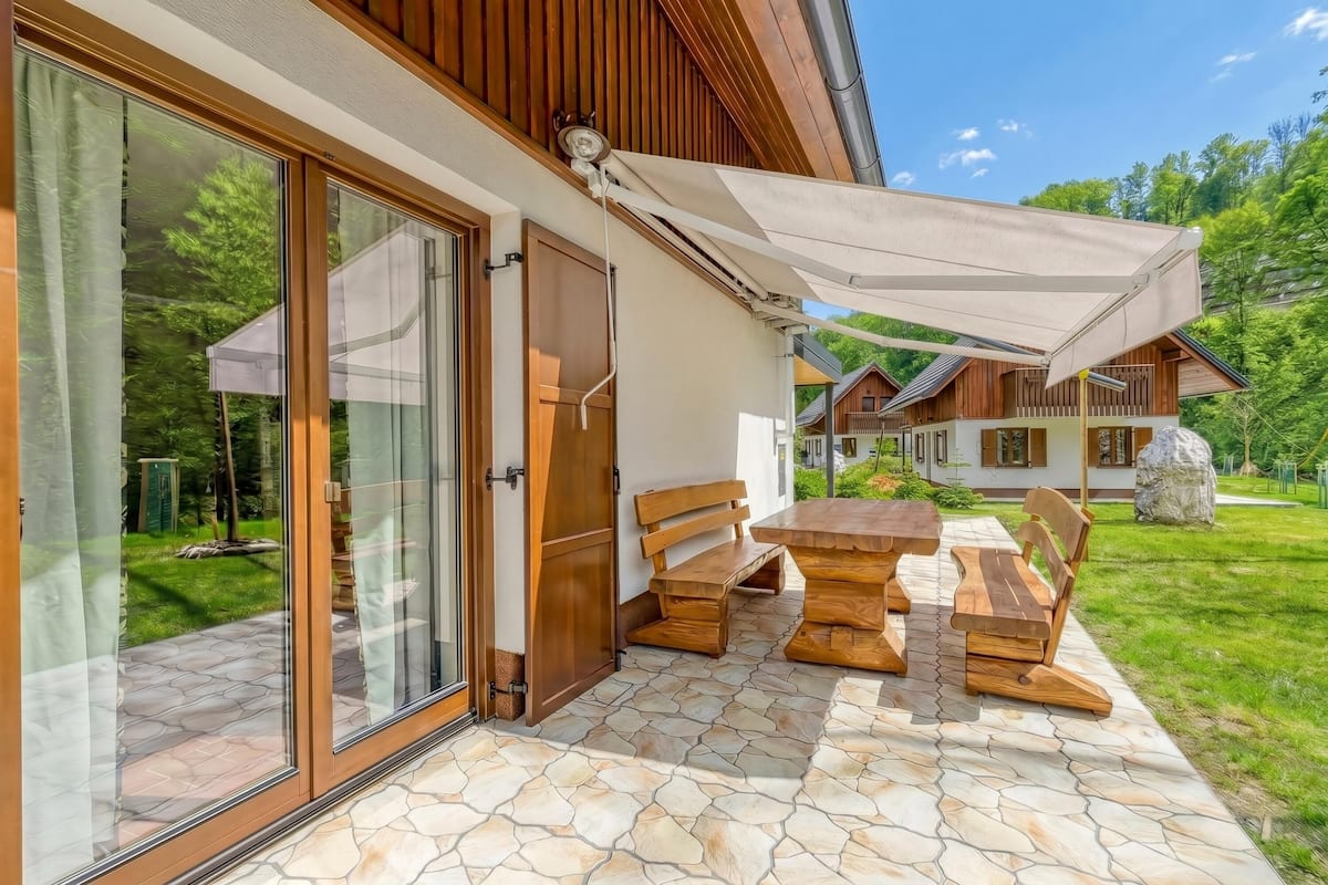 Eine Terrasse mit einem Holztisch und -bänken, ein überdachter Bereich mit einer weißen Markise und ein Boden mit Steinplatten.
