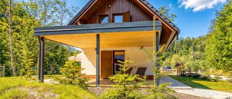 A modern wooden cabin with a covered porch, surrounded by lush greenery and a clear sky.