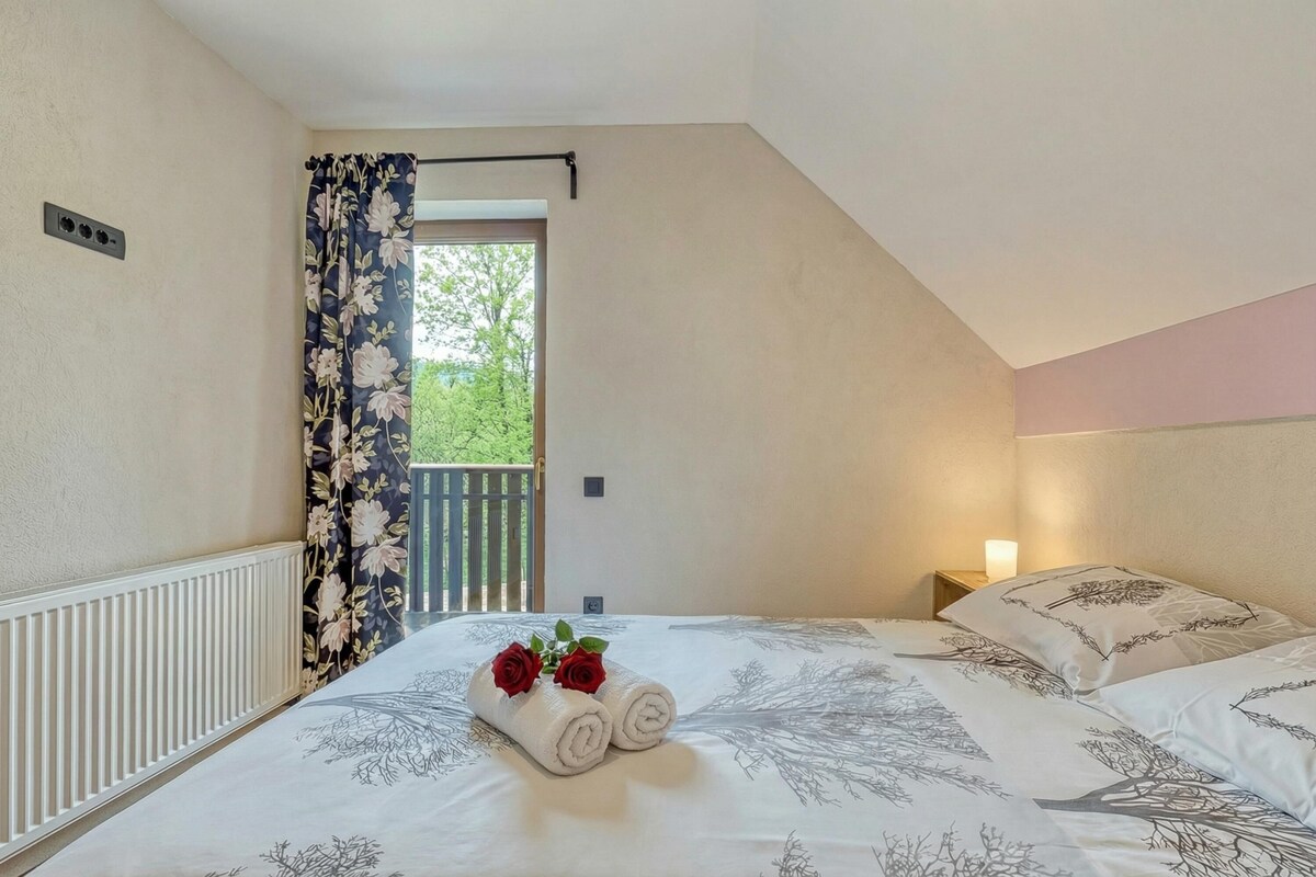 Een slaapkamer met een bed, een nachtkastje met een lamp, een raam met gordijnen en uitzicht op groen buiten.
