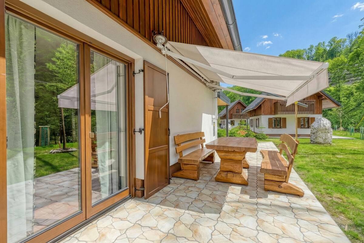 Eine Terrasse mit einem Holztisch und Bänken, ein überdachter Bereich mit einem weißen Baldachin und eine Glastür, die in einen Garten führt.