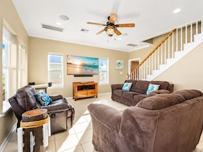 A living room with a brown leather sofa, a wooden coffee table, a ceiling fan, a TV, and a staircase. - Steps to Beach | 4BR | Deck + Ocean Views (Crystal Beach)