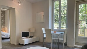 A modern, well-lit room with a round white table, a TV on a white cabinet, a dining area with chairs, and large windows offering a view of greenery.