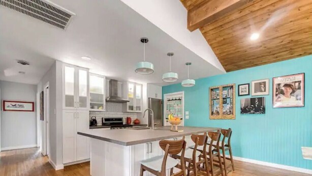 A modern kitchen with white cabinetry, a central island with bar stools, pendant lighting, and a blue accent wall with framed photos. - Mid Century Charmer, oversize fenced corner yard, fire pit,hot tub, pet friendly (Fredericksburg)