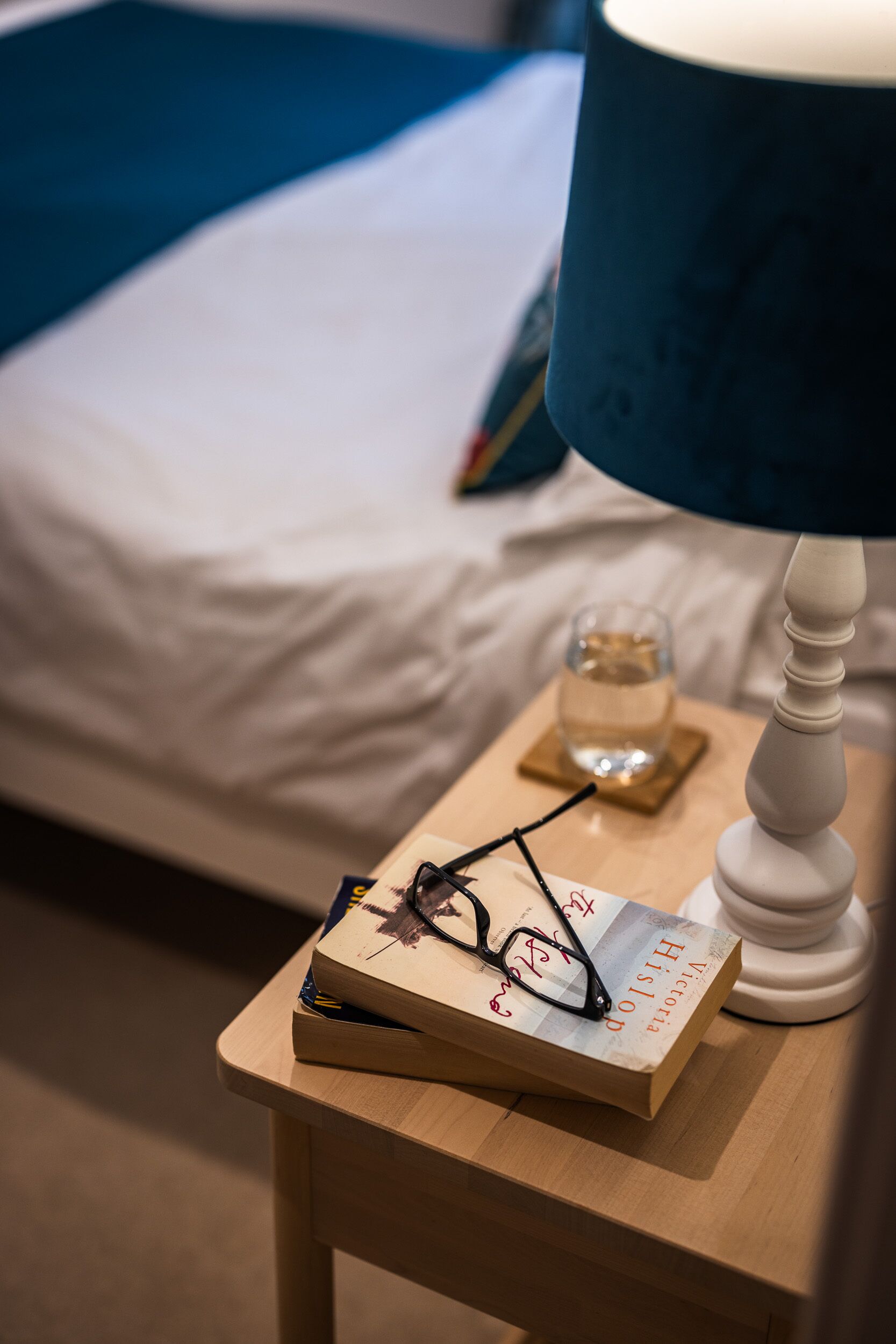 Een net opgemaakt bed met een wit laken, een nachtkastje met een lamp, een glas water en een boek met een brilretentie.