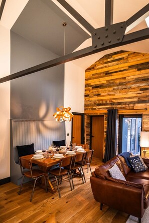 A dining area with a wooden table set for four, a brown leather sofa, and a wooden ceiling with exposed beams. - The Cow Shed @ Atlow Mill (Atlow)