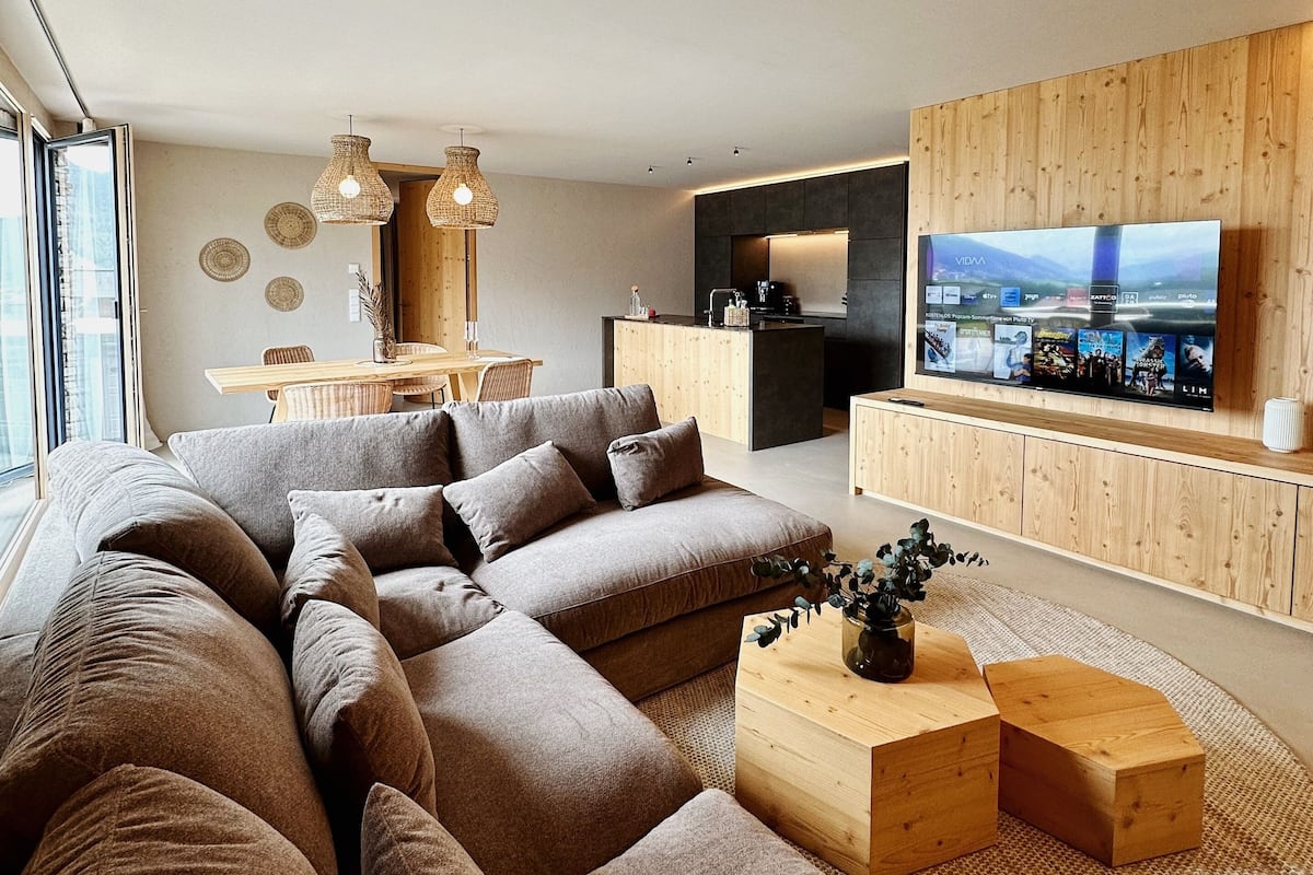 Un salon moderne avec un grand canapé d’angle, une table basse en bois et un téléviseur à écran plat fixé à un mur en bois.