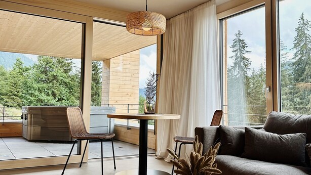 Una sala moderna con una mesa de centro de madera, un sofá y un comedor con una mesa y sillas. Grandes ventanales ofrecen vistas a árboles y montañas.